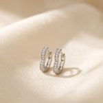 نمای نزدیک از یک جفت گوشواره زنانه حلقه‌ای ظریف و کوچک به رنگ نقره‌ای با انحنای ملایم شبیه به قلب که سطح جلویی آن با یک ردیف نگین‌های ریز و درخشان پوشیده شده است، قرار گرفته روی پس‌زمینه پارچه‌ای روشن.