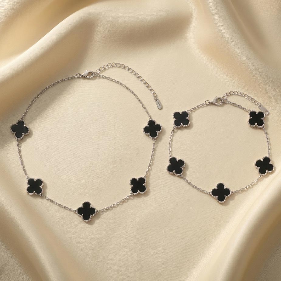 Van-Cleef-style-silver-jewelry-set-with-black-onyx تصویر یک ست دستبند و گردنبند طرح ونکلیف با زنجیر ظریف نقرهای و پلاکهای متعدد به شکل شبدر چهار برگ با رنگ مشکی که روی پارچه ساتن کرم رنگ قرار گرفتهاند.