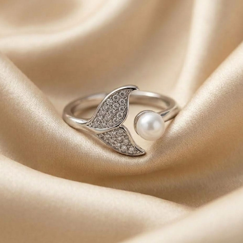 crystal-whale-tail-pearl-adjustable-ring انگشتر نقرهای با رکاب باز که یک سر آن به شکل دم وال پوشیده از نگینهای درخشان و سر دیگر آن یک مروارید کروی سفید است و روی پارچه ساتن کرم قرار دارد.