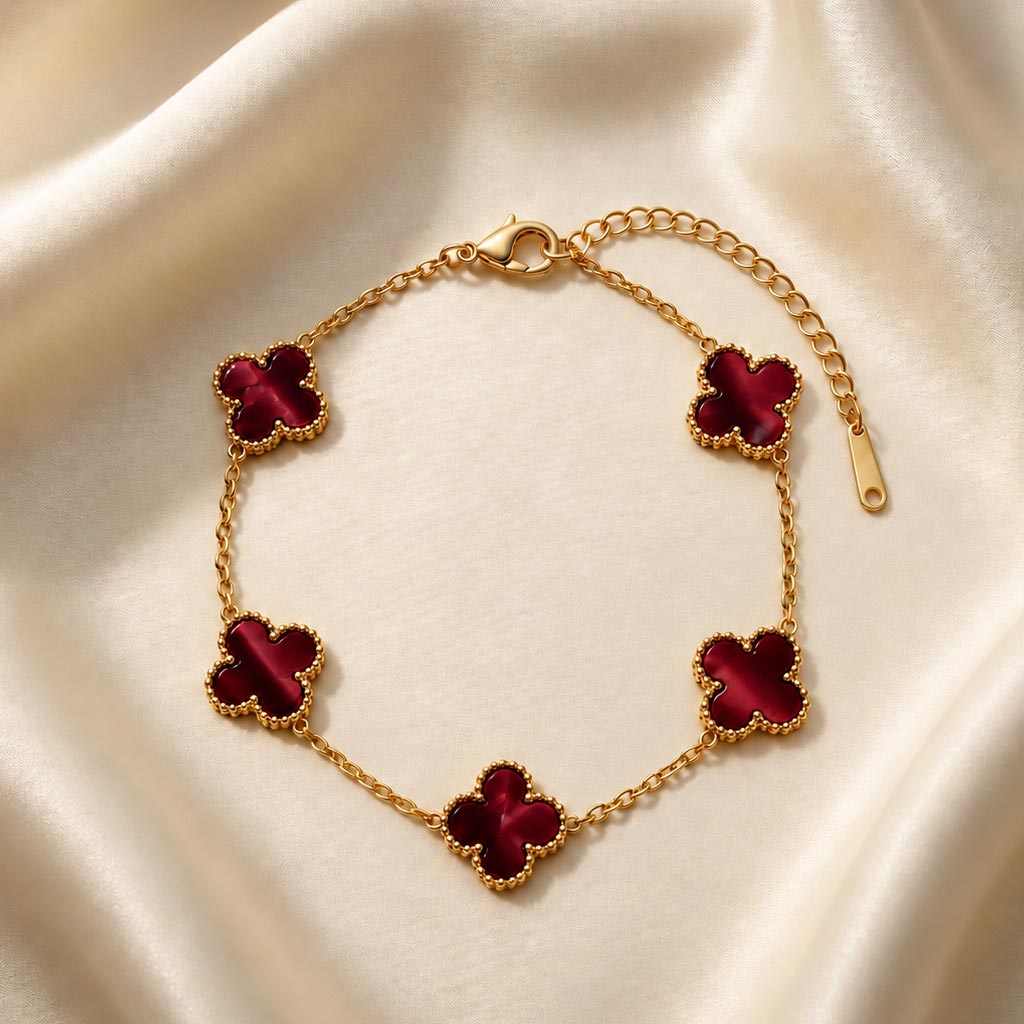 gold-van-cleef-crimson-clover-bracelet تصویر دستبند طلایی طرح ون کلیف نگین زرشکی با پنج پلاک به شکل گل شبدر که روی یک پارچه ساتن روشن قرار گرفته است.