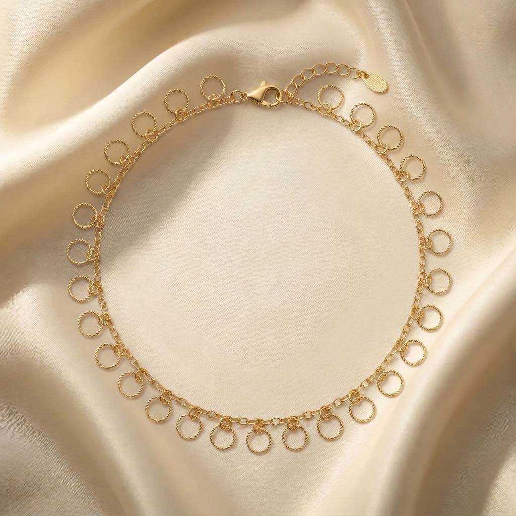 golden-circle-dangle-anklet-twisted پابند زنانه به رنگ طلایی شامل یک زنجیر اصلی ظریف و آویزهای متعدد به شکل حلقههای دایرهای کوچک با طراحی بافتدار (طنابی)، دارای قفل طوطی و زنجیر تنظیم سایز که روی پسزمینه پارچه ساتن کرمرنگ قرار گرفته است.