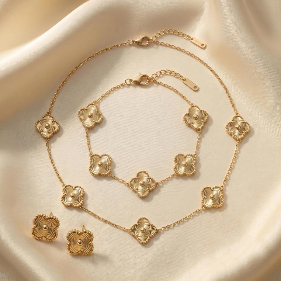 golden-van-cleef-sunburst-3-piece-set ست سه تکه زیورآلات طلایی رنگ شامل یک گردنبند با پنج پلاک شبدر، یک دستبند با پنج پلاک و یک جفت گوشواره میخی. سطح پلاکهای این ست ۳ تکه طرح ون کلیف طلایی دارای خطوط شیاردار و بافت خورشیدی است که روی یک پارچه ساتن کرم رنگ قرار گرفتهاند.
