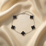 دستبند طرح ون کلیف نقره ای قرار گرفته روی پارچه ساتن با پنج پلاک شبدر چهار برگ که داخل آن‌ها مشکی است و حاشیه نقطه‌کوب شده دارند.