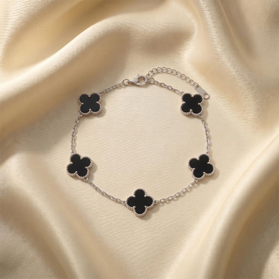 silver-bracelet-van-cleef-five-black-clover دستبند طرح ون کلیف نقره ای قرار گرفته روی پارچه ساتن با پنج پلاک شبدر چهار برگ که داخل آنها مشکی است و حاشیه نقطهکوب شده دارند.