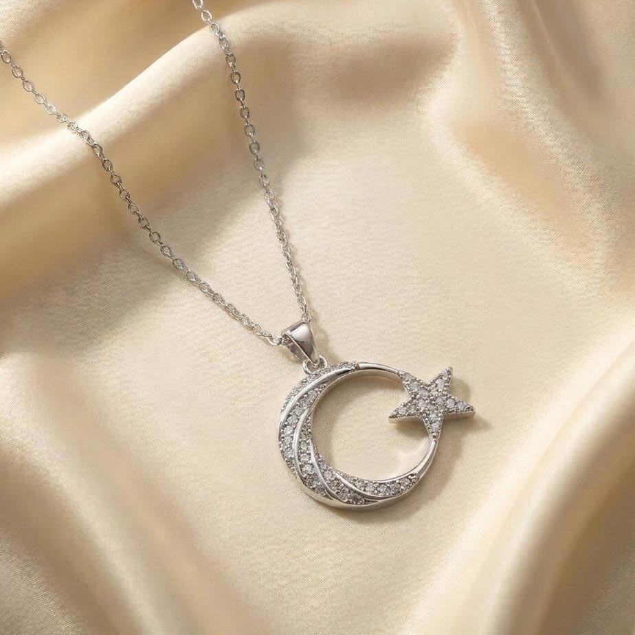 silver-crescent-moon-star-crystal-necklace گردنبند طرح ماه و ستاره نقرهای با پلاکی به شکل هلال ماه شیاردار و یک ستاره پنجپر متصل به آن که هر دو با نگینهای ریز درخشان پوشانده شدهاند، رها شده روی پارچه ابریشمی کرم رنگ.