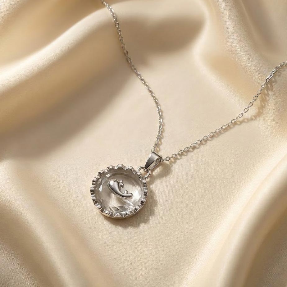 silver-dolphin-crystal-round-necklace گردنبند طرح وال نقرهای با پلاک قابدار دایرهای که در مرکز آن یک شیشه شفاف تراشخورده و روی آن یک بچه وال براق قرار دارد، رها شده روی پارچه ابریشمی کرم رنگ.