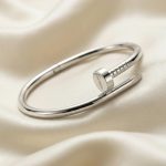 یک دستبند استیل براق طرح میخ برند کارتیه به رنگ نقره‌ای (سیلور) با حکاکی ظریف نام Cartier روی سر میخ، که روی پس‌زمینه پارچه ساتن کرم/سفید رنگ قرار گرفته است.