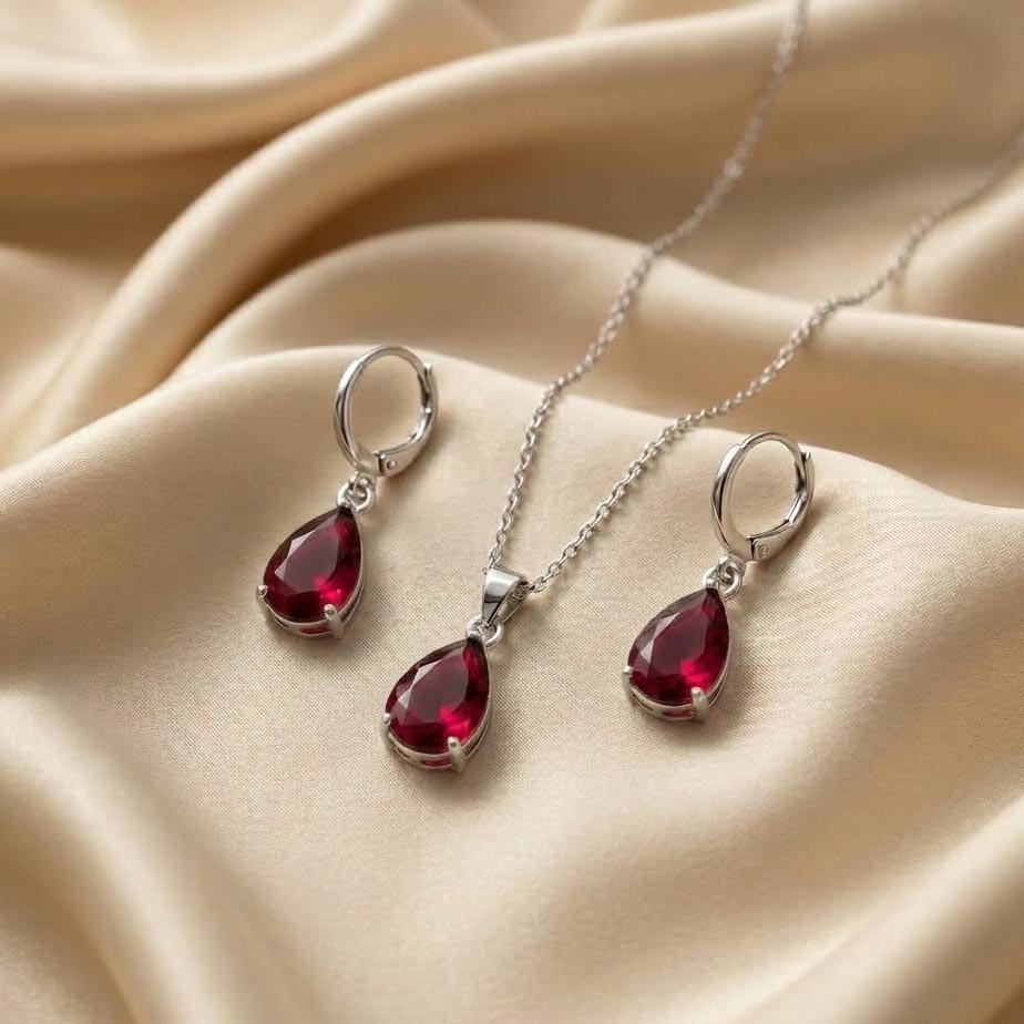 silver-teardrop-jewelry-set-red-stone تصویر یک نیم ست طرح اشکی نقرهای شامل یک گردنبند ظریف و گوشوارههای آویز قفلی که دارای نگینهای بزرگ تراشخورده به رنگ قرمز (طرح یاقوت) هستند و روی پارچه ساتن کرم قرار دارند.