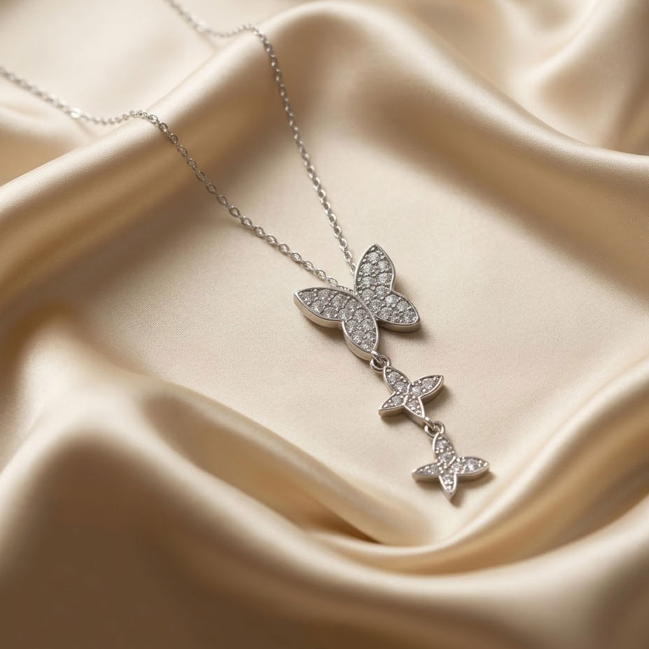 silver-triple-butterfly-drop-necklace یک گردنبند نقرهای از کالکشن پروانه شامل یک پروانه بزرگ نگیندار که دو پروانه کوچکتر به صورت دنبالهدار به زیر آن متصل شدهاند و روی پارچه ابریشمی کرم رنگ قرار دارد.