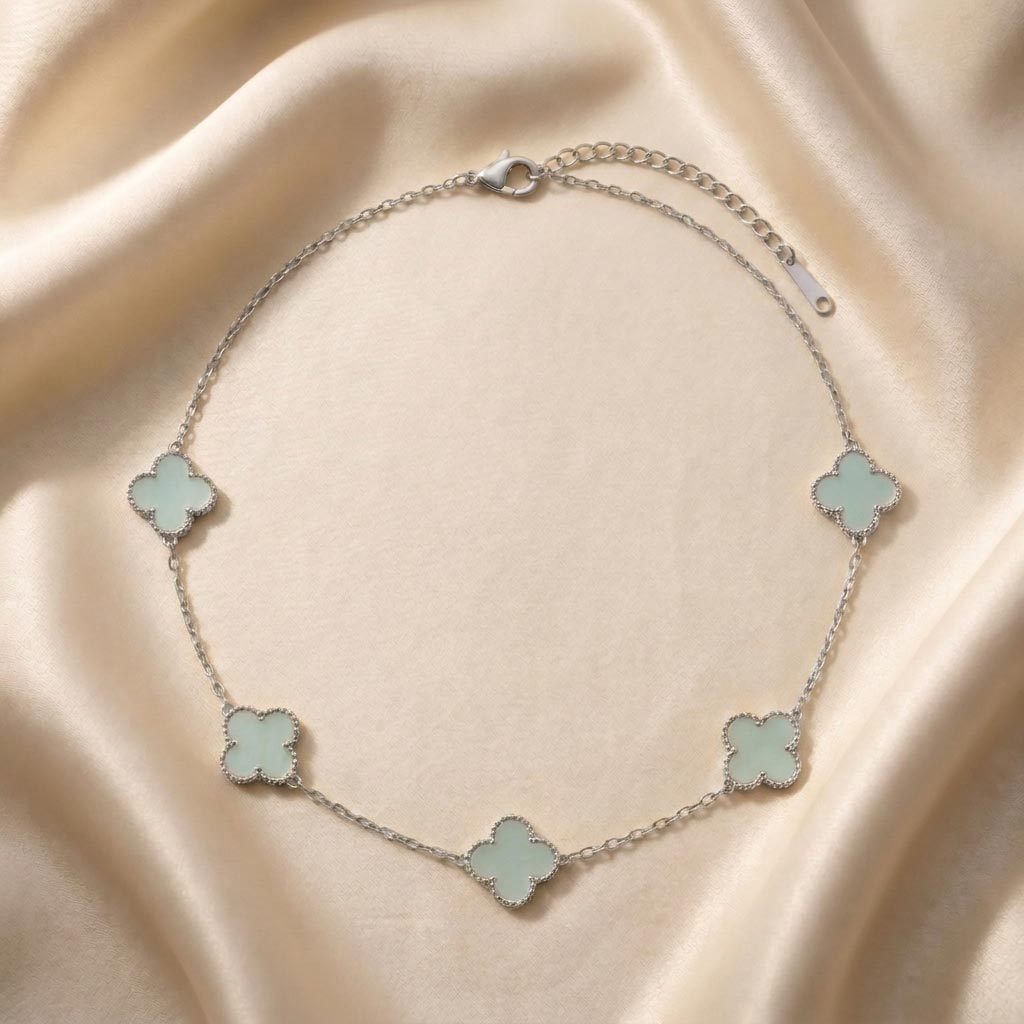 silver-van-cleef-clover-necklace-tiffany-blue گردنبند نقرهای رنگ با پنج پلاک ظریف به شکل شبدر چهار برگ یا طرح ون کلیف با رنگ داخلی آبی تیفانی که روی یک پارچه ابریشمی کرمرنگ قرار گرفته است، گردنبند طرح ون کلیف نقرهای.