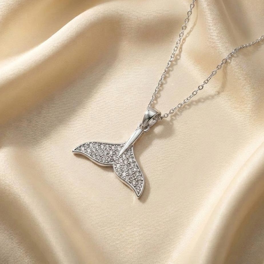silver-whale-tail-necklace-with-crystals-with-jewel یک گردنبند ظریف با پلاک طرح دم وال نقرهای نگیندار که روی پارچه ساتن طلایی قرار دارد، نمایش زیبایی گردنبند طرح دم وال نقرهای.