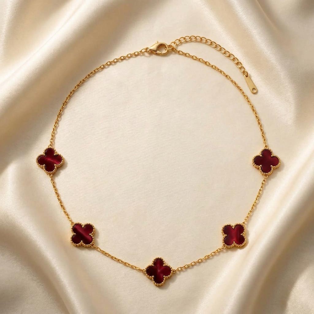 van-cleef-clover-necklace-burgundy گردنبند طلایی رنگ با پنج پلاک ظریف به شکل شبدر چهار برگ یا طرح ون کلیف با رنگ داخلی زرشکی عمیق که روی یک پارچه ابریشمی کرمرنگ قرار گرفته است، گردنبند طرح ون کلیف طلایی.