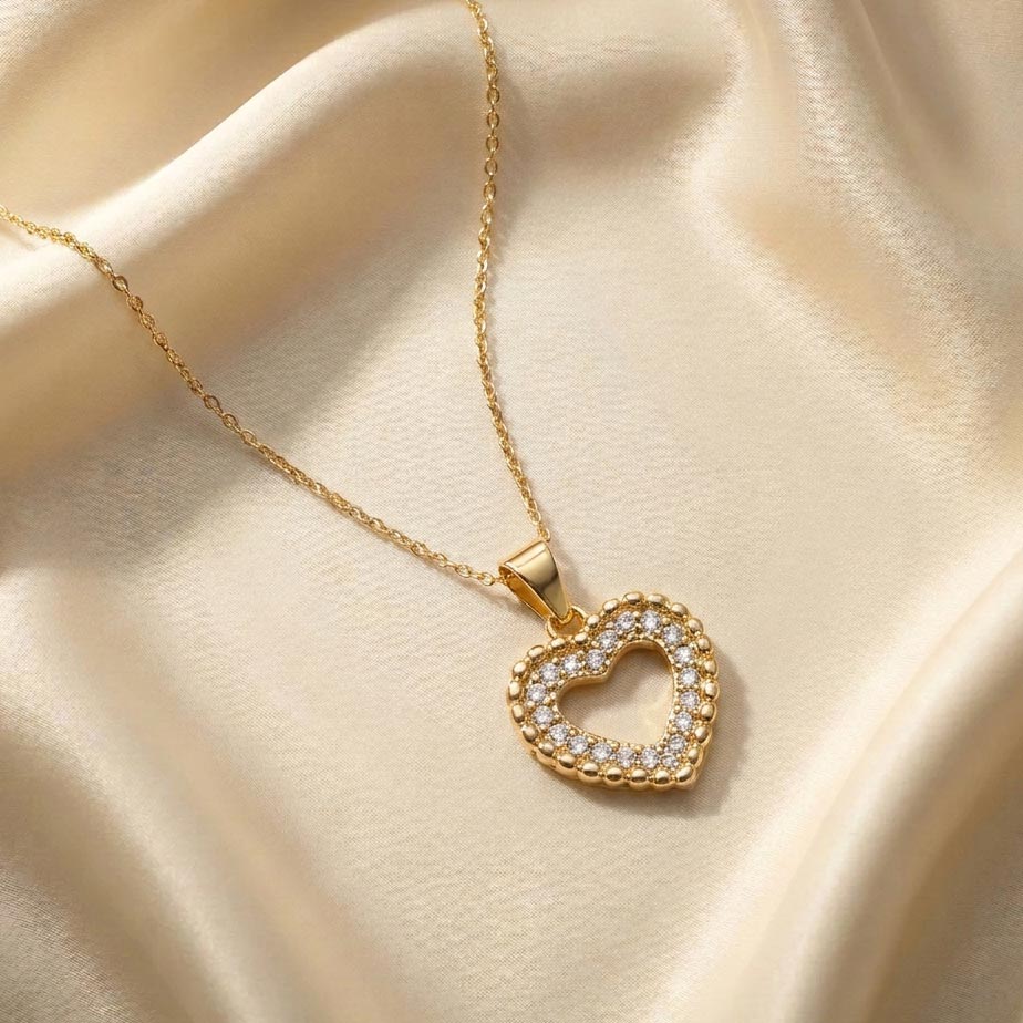 womens-gold-open-heart-crystal-necklace گردنبند طلایی رنگ با پلاک قلب توخالی که حاشیه آن با نگینهای ریز سفید و نقطههای برجسته طلایی تزئین شده و روی پارچه ساتن کرم رنگ قرار دارد، نمایشی از زیبایی گردنبند طلایی با پلاک قلب.