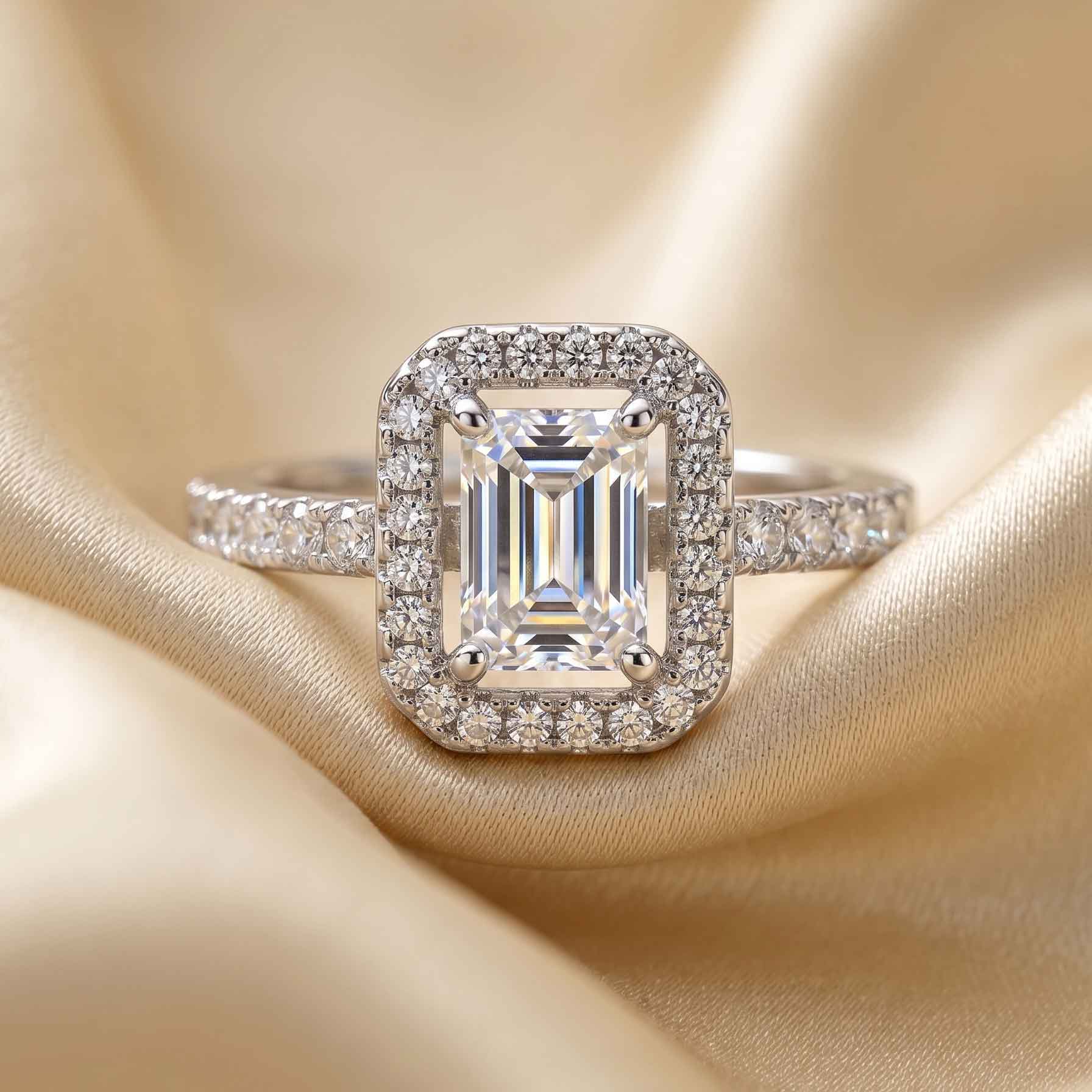 Elora-halo-solitaire-ring یک انگشتر نقرهای رنگ روی پارچه ساتن طلایی که یک نگین بزرگ مستطیلی در وسط و نگینهای ریز در اطراف و روی رکاب آن قرار دارد و درخشش انگشتر طرح جواهر را نشان میدهد.
