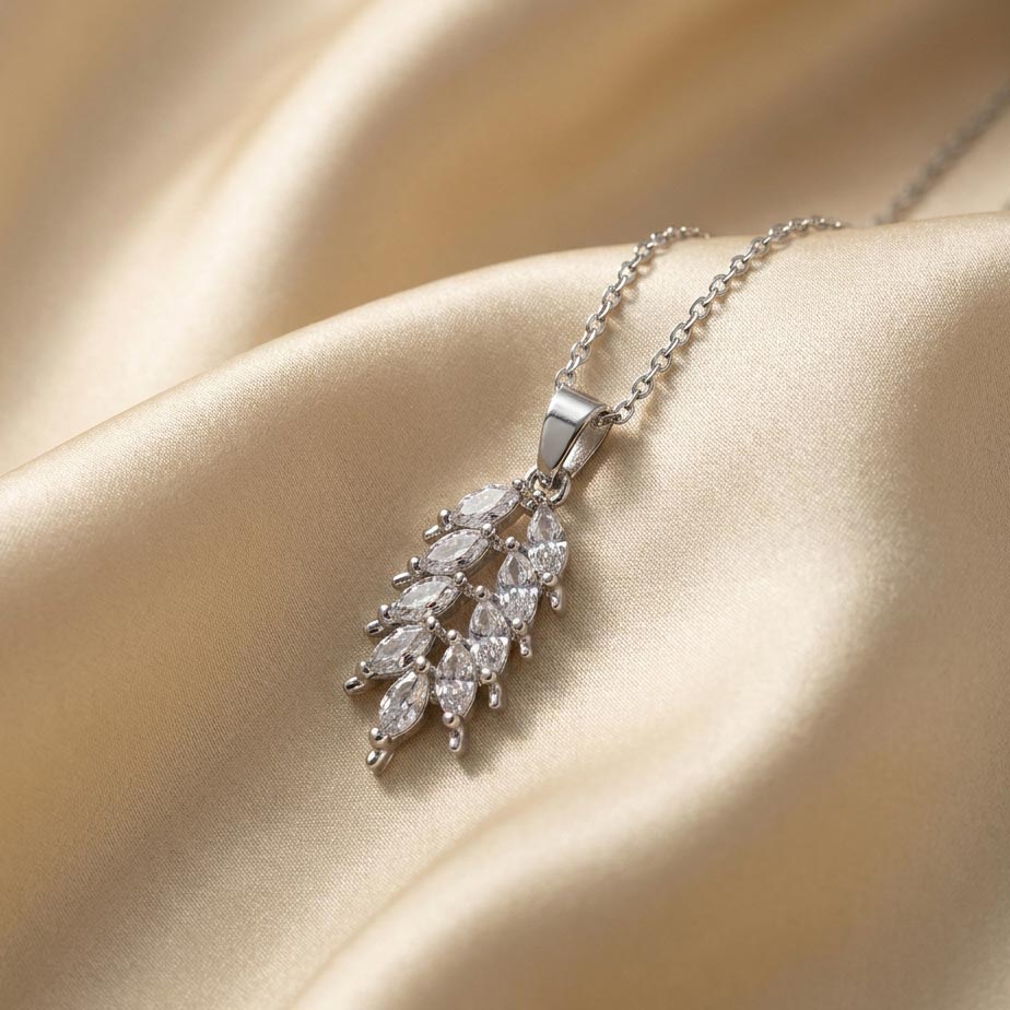 womens-silver-leaf-crystal-necklace گردنبند نقرهای رنگ با پلاک طرح برگ تشکیل شده از نگینهای مارکیز روی پارچه ساتن کرم که درخشش گردنبند طرح برگ نگیندار را نشان میدهد.